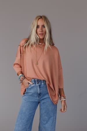 A woman with long blonde hair wears The Nest Cozy Cool Oversized Tee in Coral and high-waisted blue jeans, accessorized with layered necklaces and bracelets. She poses with one hand in her pocket against a plain gray background.