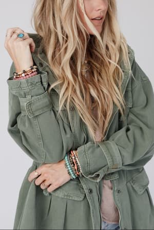 A woman with long, wavy blonde hair wears the Layers On Layers Perfect Boho Parka in olive, paired with colorful beaded bracelets and rings. She stands with her arms crossed, gazing slightly down and to the side.