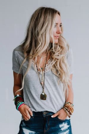 A woman with long, wavy blonde hair wears the Scoop Neck Bralette Tee - Gray, layered necklaces, and stacked bracelets. She looks to the side with one hand in her ripped blue jeans pocket against a plain light background.