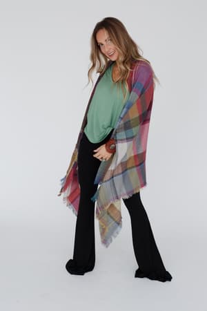 A woman with long, wavy hair stands smiling against a white background, wearing a green top, black pants, and the Cozy Plaid Pocket Ruana Wrap in Fuchsia draped over her shoulders, posing with one knee slightly bent.