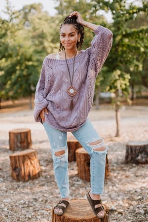 A woman poses outdoors on a tree stump, wearing the Golden Hour Bubble Sleeve Sweater Dress in lavender. She stands confidently among trees and cut logs, accessorized with braided hair and a long necklace.