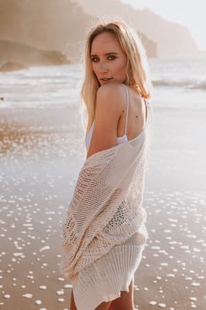 A woman with long blonde hair stands on the beach at sunset, wearing the Brenden Knit Netted Cardigan in Ivory over a white swimsuit. She looks over her shoulder at the camera as the sun casts a warm glow on the waves behind her.