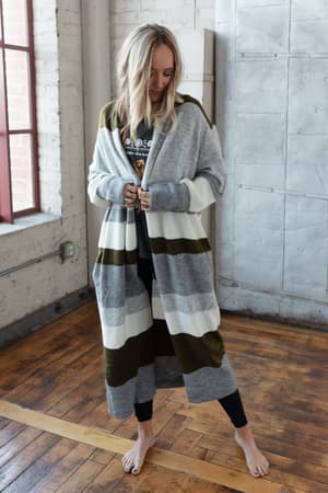 A barefoot woman indoors wears black leggings, a printed T-shirt, and The Nest So Cozy Oversized Striped Knit Duster in Olive Multi with wide gray, white, and olive green stripes. She looks down and touches the duster.