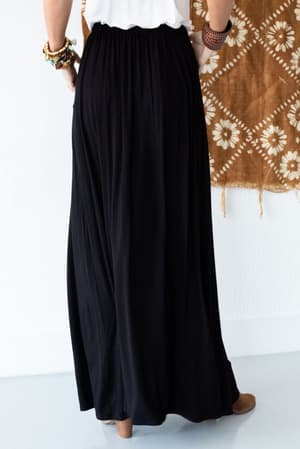 A person stands facing away, wearing The Perfect Pocketed Maxi Skirt - Black, a white top, brown shoes, and colorful bracelets. A brown wall hanging with white floral patterns appears in the background.