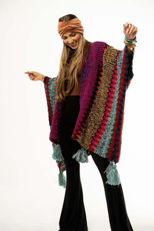 A woman wearing the vibrant Desert Dreams Crochet Ruana - Magenta, a tan headband, and black pants stands with arms raised and smiles against a white background—a must-have for every boho closet.