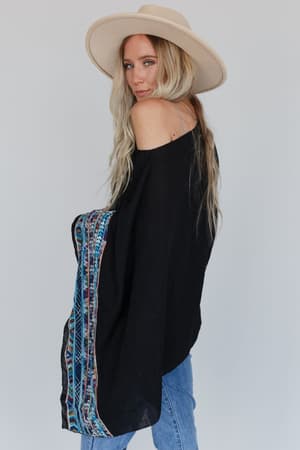 A woman with long blonde hair wears a wide-brimmed beige hat, blue jeans, and the Fallon Embroidered Sleeve Poncho in black, standing against a plain light background.