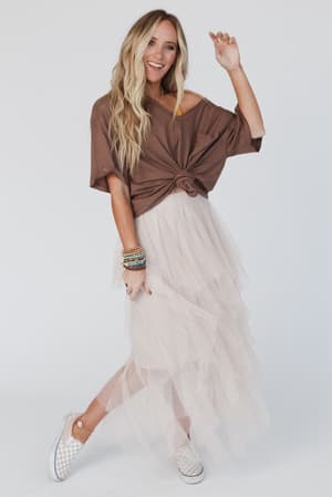 A woman with long blonde hair smiles and poses with one arm raised, wearing a brown knotted t-shirt, the Boho Ballerina Skirt - Nude with a handkerchief hemline, checkered slip-on sneakers, and colorful bracelets against a white background.