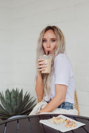 A woman with long blonde hair sips an iced coffee at an outdoor table, wearing the Scoop Neck Bralette Tee in white. She looks at the camera while food and a plant are visible beside her.