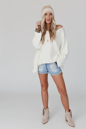 A woman wears the Dana Oversized Diamond Texture Sweater in ivory off one shoulder, paired with light denim shorts, a beige beanie, tan ankle boots, layered necklaces, and bracelets while posing against a plain white background.