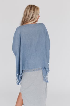 A woman with long blonde hair, shown from the back, wears the Fallon Embroidered Sleeve Tassel Poncho in blue over a light gray dress with a side slit. The background is plain white.