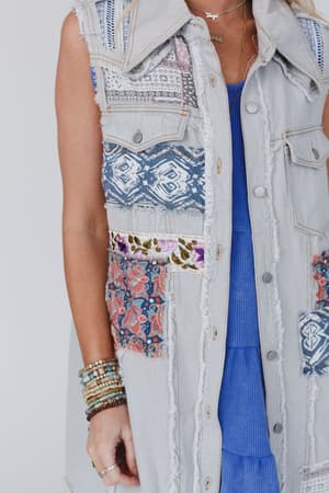 She wears the One Of A Kind Sleeveless Denim Dress Vest - Denim Multi, featuring colorful patchwork, frayed edges, and metal buttons over a blue dress. Layered bracelets and a necklace add to her bold, boho-inspired style.