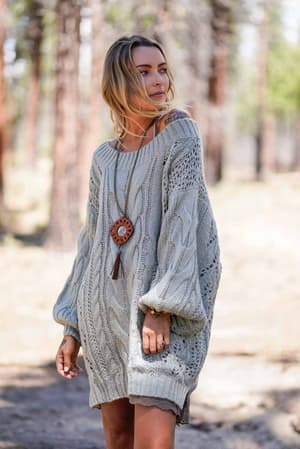 A woman stands outdoors in a forest, wearing the Golden Hour Bubble Sleeve Sweater Dress in sage and accessorized with a long pendant necklace.
