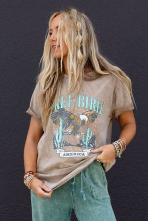 A woman with long blonde hair wears a Fly Free Graphic Tee - Mocha featuring an eagle and cacti, paired with green pants and beaded bracelets. She stands against a dark wall, smiling as she looks to the side.