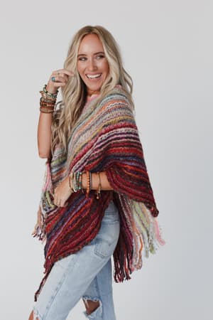 A smiling woman with long blonde hair wears the Madeleine Poncho - Multi with ripped light blue jeans, accessorized with stacked bracelets and rings, standing against a plain light background.