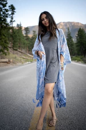 A woman with long dark hair stands on a mountain road, looking relaxed at the camera. She wears a grey dress, sandals, and the Crushin' On You Velvet Kimono in Light Blue.