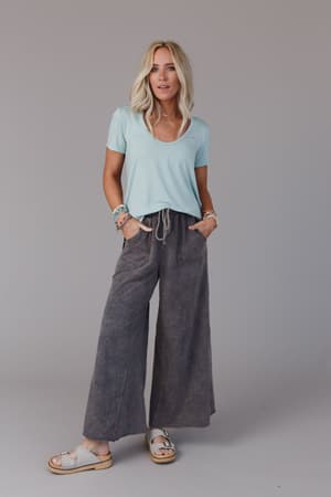 A woman with blonde hair wears the Scoop Neck Bralette Tee in Washed Seafoam, gray wide-leg drawstring pants, sandals, and bracelets while standing against a plain gray background.