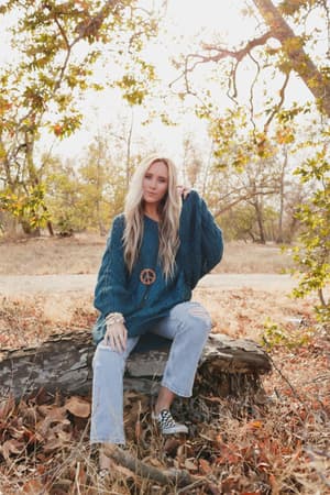 A woman with long blonde hair sits on a fallen tree in a dry, autumn forest, wearing the Golden Hour Bubble Sleeve Sweater Dress in teal. Sunlight filters through the trees behind her.