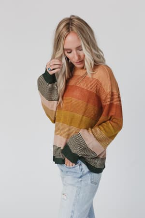 A woman with long blonde hair wears the Going Places Gradient Stripe Sweater - Mustard, featuring oversized gradient stripes of orange, yellow, and green. She pairs it with light blue jeans and stands against a plain background, looking down.