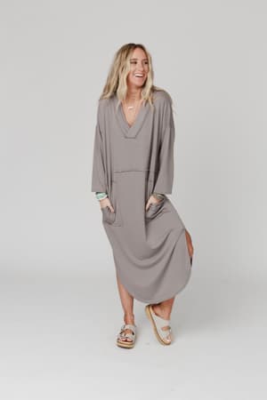 A woman smiles with her hands in the pockets of The Nest Meadow Long Sleeve Slit Maxi Dress - Coco. She pairs it with slide sandals, layered jewelry, and wears her long blonde hair down. The background is plain and light-colored.
