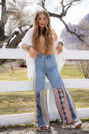 A woman with long wavy hair stands by a white fence outdoors, wearing a tan top, white fringed shawl, layered bracelets, sandals, and Magnolia Patchwork Jeans - Washed Denim. Trees and mountains create a scenic backdrop.