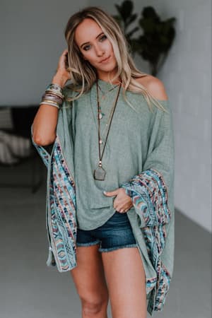 A woman with long blonde hair wears the Fallon Embroidered Sleeve Poncho in sage, paired with denim shorts, layered necklaces, and multiple bracelets. She stands indoors, facing the camera, one hand touching her hair.