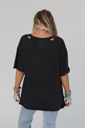 A person with long blonde hair stands with their back to the camera, wearing the Share The Love Graphic Tee in charcoal, light blue jeans, and multiple colorful bracelets on both wrists.