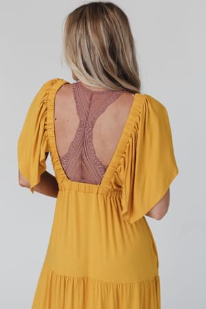 A woman with blonde hair faces away, wearing a yellow stretch knit dress with ruched sleeves and an open back that reveals the Evermore Seamless Lace Racerback Bralette in mauve, set against a plain, light-colored background.