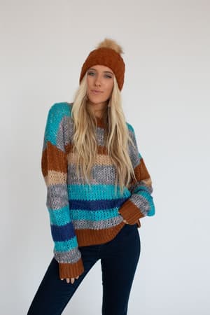 A woman with long blonde hair wears the Go For It Striped Sweater - Blue, a chunky knit in blue, brown, and gray stripes, paired with dark jeans and a brown beanie. She poses against a white background, looking at the camera.