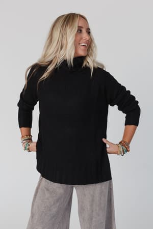 A woman with long blonde hair smiles and looks to the side against a plain light background, wearing the Danny Sweater in black, loose gray pants, and colorful beaded bracelets.