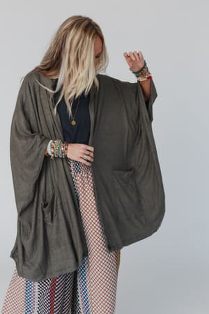 A woman with long blonde hair wears The Nest Boho Essential Hooded Ribbed Sweater in olive, a black top, patterned pants, and beaded bracelets. She stands against a plain light background, looking down with one arm bent.