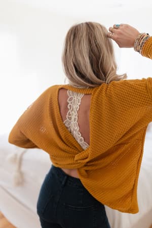 A blonde woman stands with her back to the camera, wearing The Nest Flattering Open Back Thermal Knit Top in Mustard, revealing a white lace bralette and stacked bracelets, all in a softly lit room.