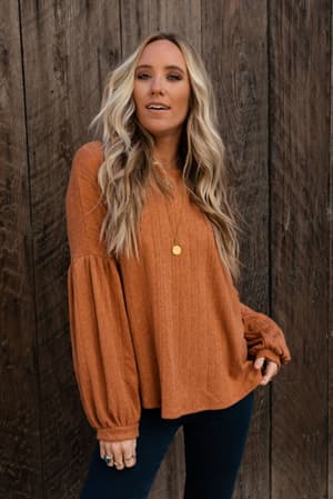 A woman with long, wavy blonde hair stands by a wooden wall wearing The Nest Sweet Heart Balloon Sleeve Knit Sweater in Burnt Orange and dark jeans, accessorized with a gold necklace and rings, one hand on her hip.