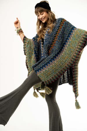 A woman smiles playfully, wearing the Desert Dreams Crochet Ruana - Cobalt with tassels, gray flared pants, a black headband, and bracelets for a boho vibe against a white background.