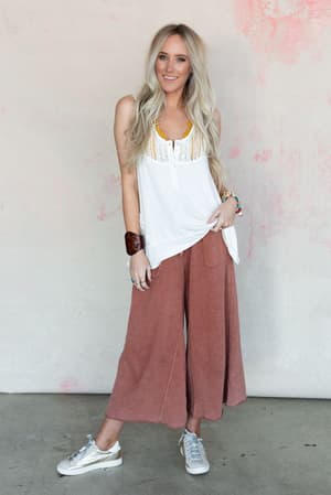 A woman with long blonde hair smiles against a pink and white wall, wearing a sleeveless white top, the Relaxing Robin Wide Leg Pant in Red Bean, white sneakers, and colorful jewelry.