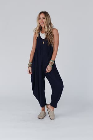 A woman with long blonde hair smiles as she stands against a plain light gray background, wearing The Perfect Harem Jumpsuit - Navy over a white top with beige slip-on shoes and bracelets on both wrists.