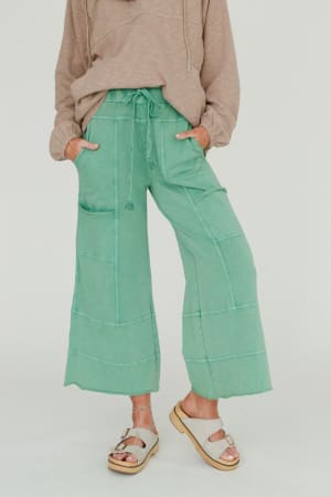 A person stands with hands in pockets, wearing the Lucky One Wide Leg Pant in green, styled with a light brown long-sleeve hoodie and white platform sandals. The relaxed boho look is set against a plain white background.