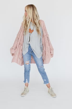 A woman with long blonde hair stands against a white background, wearing the Cara Oversized Cardigan in pink over a gray top, ripped blue jeans, and beige sneakers, accessorized with a butterfly pendant necklace.