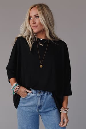 A woman with long blonde hair wears The Nest Cozy Cool Oversized Tee in black and light blue jeans, accessorized with layered necklaces and colorful bracelets, standing against a plain gray background.