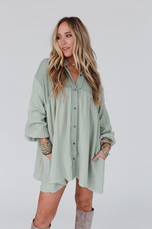 A woman with long, wavy blonde hair poses in the Sunday Kind Of Love Dress - Sage, accessorized with bracelets and tall gray boots, against a plain white background.