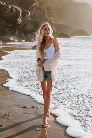 A woman with long blonde hair walks barefoot along the beach in a white tank top, denim shorts, and the Brenden Knit Netted Cardigan - Ivory. Waves wash over the sand, and rocky cliffs rise in the background.