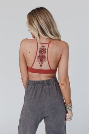 A woman with long blonde hair stands with her back to the camera, wearing the Tattoo Bralette - Rust and gray pants. An intricate floral tattoo is visible on her upper back, and she wears multiple beaded bracelets on her left wrist.