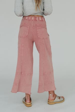 A person faces away, wearing the Lucky One Wide Leg Pant in mauve knit with two back pockets, paired with a striped long-sleeve top and beige sandals. The background is plain and light-colored.