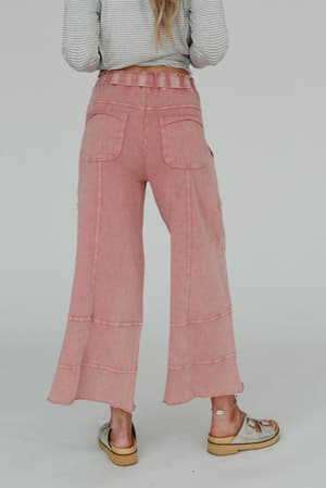 A person faces away, wearing the Lucky One Wide Leg Pant in mauve knit with two back pockets, paired with a striped long-sleeve top and beige sandals. The background is plain and light-colored.