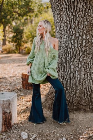 A woman with long blonde hair stands outdoors by a large tree, wearing the Golden Hour Bubble Sleeve Sweater Dress in light green. The natural background features scattered leaves, tree stumps, and greenery.