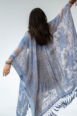 A person with long, dark hair is seen from behind, wearing the Crushin' On You Velvet Kimono in light blue. Their arms are raised, showing off the kimono’s flowing fabric and elegant design.