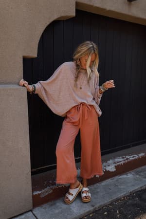 A woman with long blonde hair in loose braids wears The Nest Fly With Me Batwing Sleeve Knit Sweater in Sand, paired with wide-legged rust-colored pants, white sandals, and bracelets, standing by a dark wooden door and concrete wall.