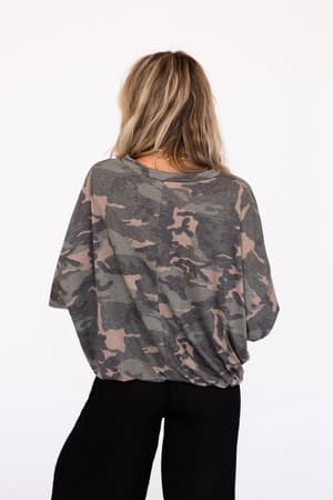 A person with long, wavy blonde hair is seen from behind wearing the Here For Camo Top - Olive, featuring an oversized slouchy fit, paired with black pants against a plain white background.