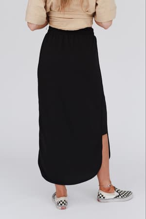 Someone stands with their back to the camera in a beige top and The Nest So Comfy Drawstring Maxi Skirt - Black, featuring a side slit and elastic waist, paired with black-and-white checkerboard slip-on shoes against a plain light background.