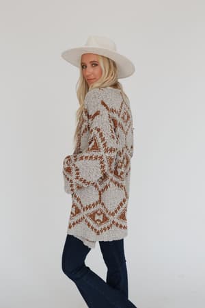 A woman wearing the Free to Roam Cardigan in gray, styled with a wide-brimmed white hat and dark jeans, stands against a plain white background, glancing over her shoulder at the camera.