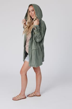 A woman with long blonde hair smiles sideways, wearing the Layers On Layers Perfect Boho Parka in olive over a light pink top, denim shorts, and tan sandals. She holds her hood with both hands against a plain white background.